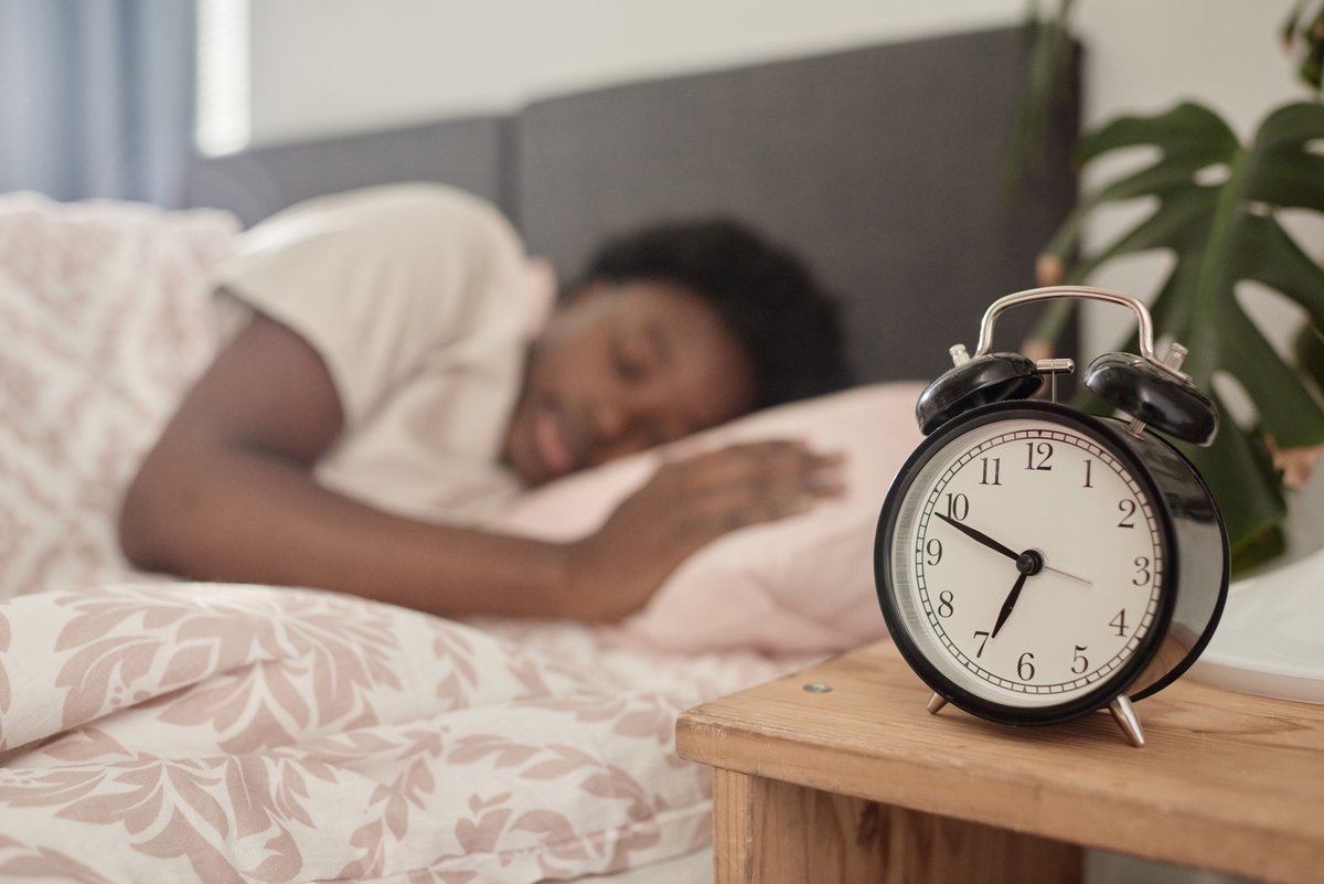 Despertador na mesa de cabeceira, com uma mulher a dormir ao fundo