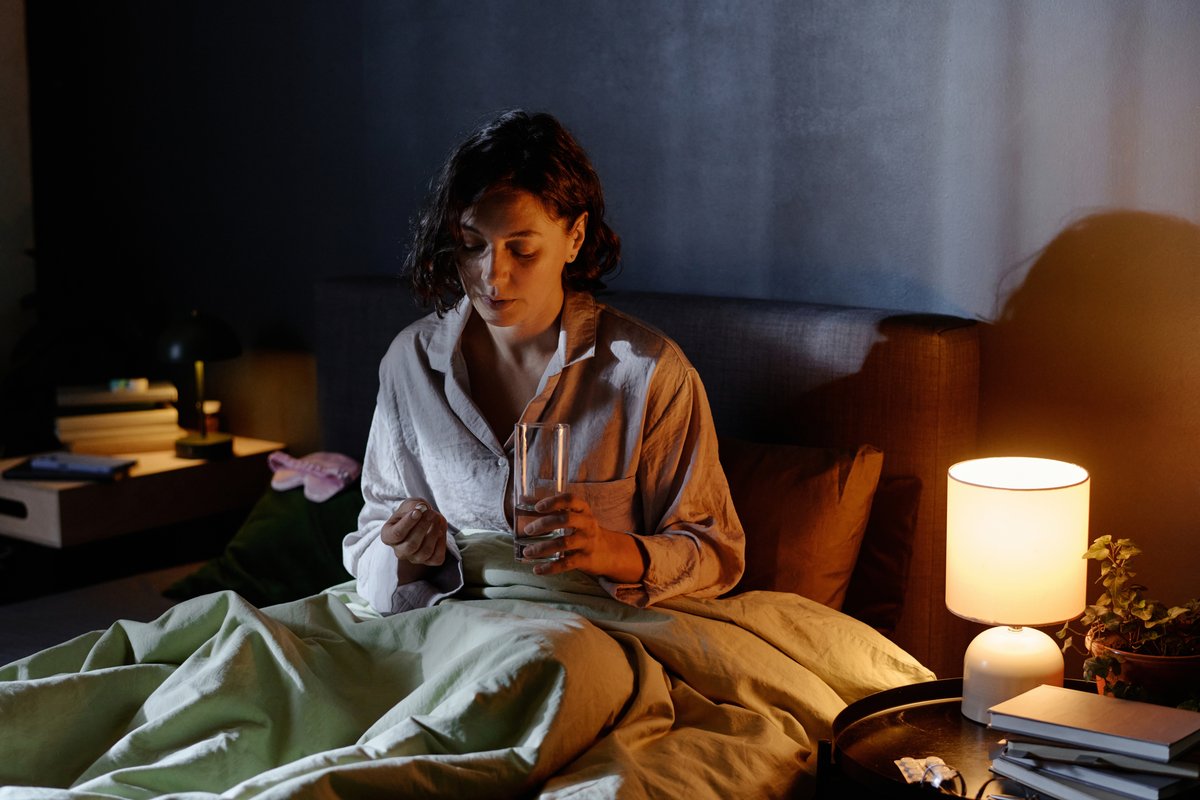 Mulher sentada na cama, de noite, a segurar um comprimido e um copo de água